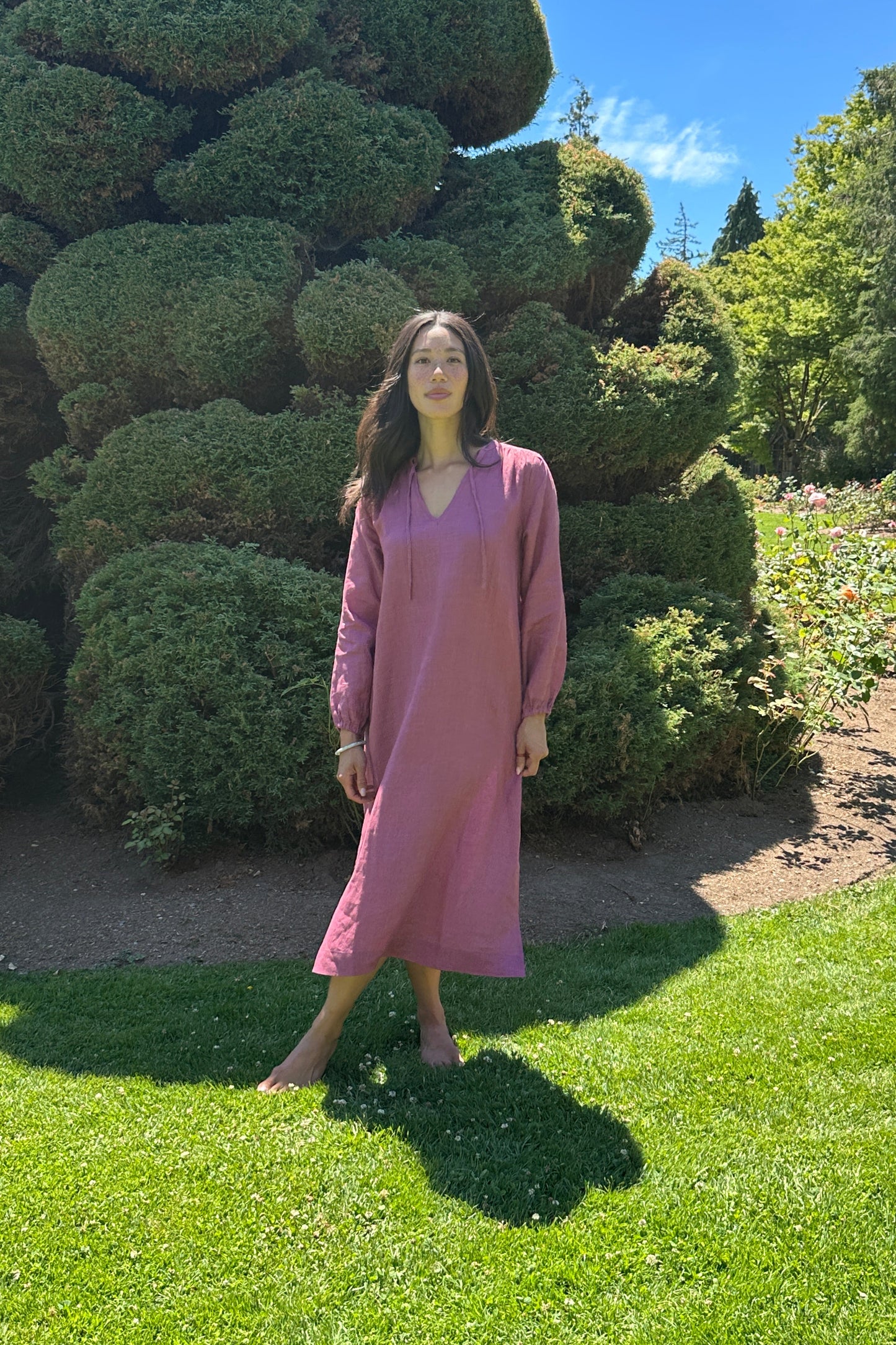 A woman wearing a Divine Rose long-sleeve maxi dress stands on a garden path between manicured green bushes and a grassy lawn under a blue sky.